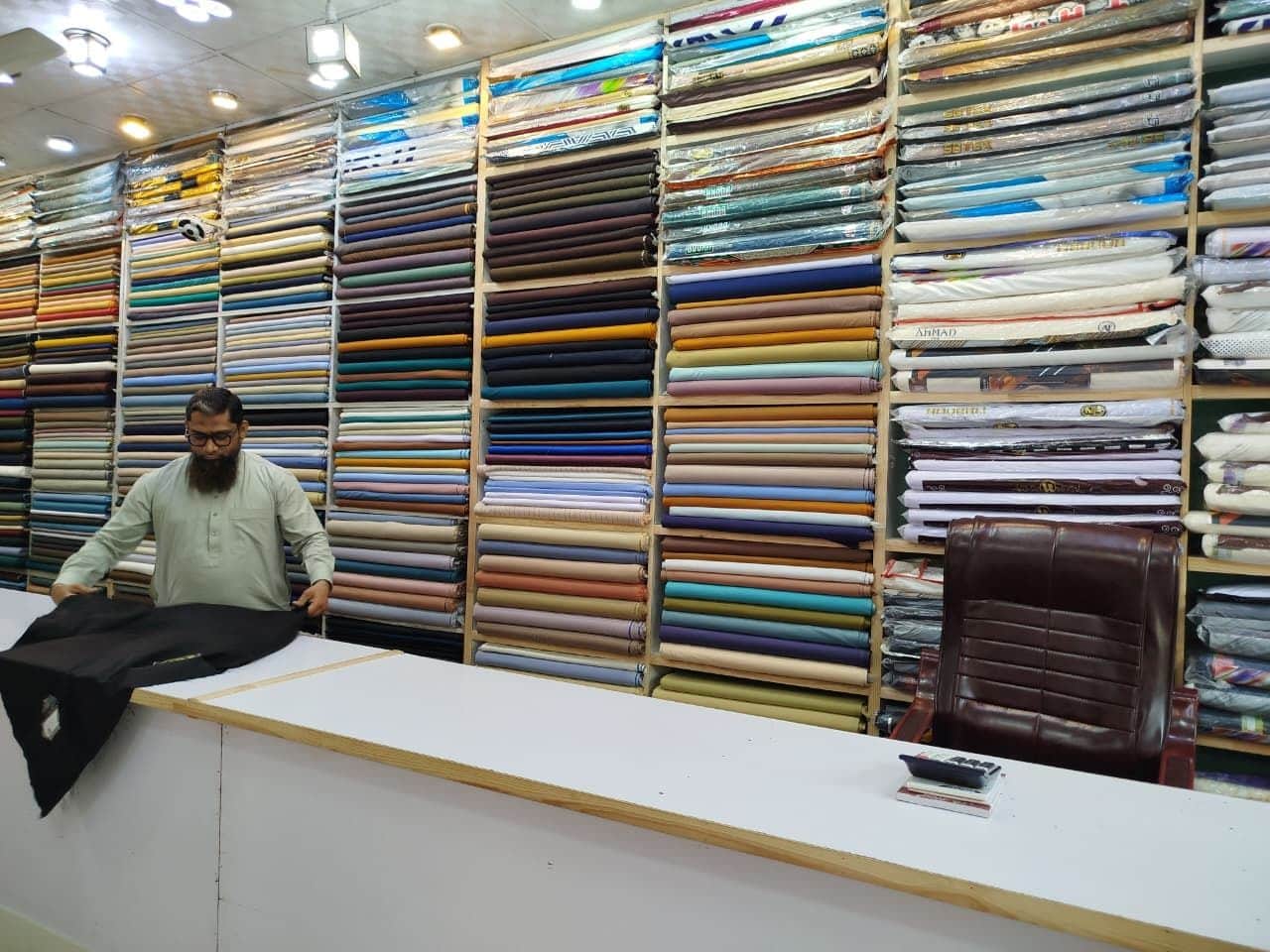 Opening a length of black suiting at the counter, in front of shelves of gents wash & wear at Shahzad Cloth House